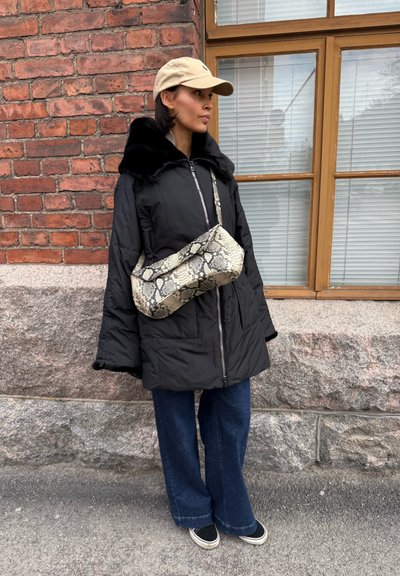 Mujer con gorra beige, abrigo negro con cuello de piel, jeans azules anchos y bolso de estampado de serpiente, de pie junto a una pared de ladrillo y piedra con una ventana.