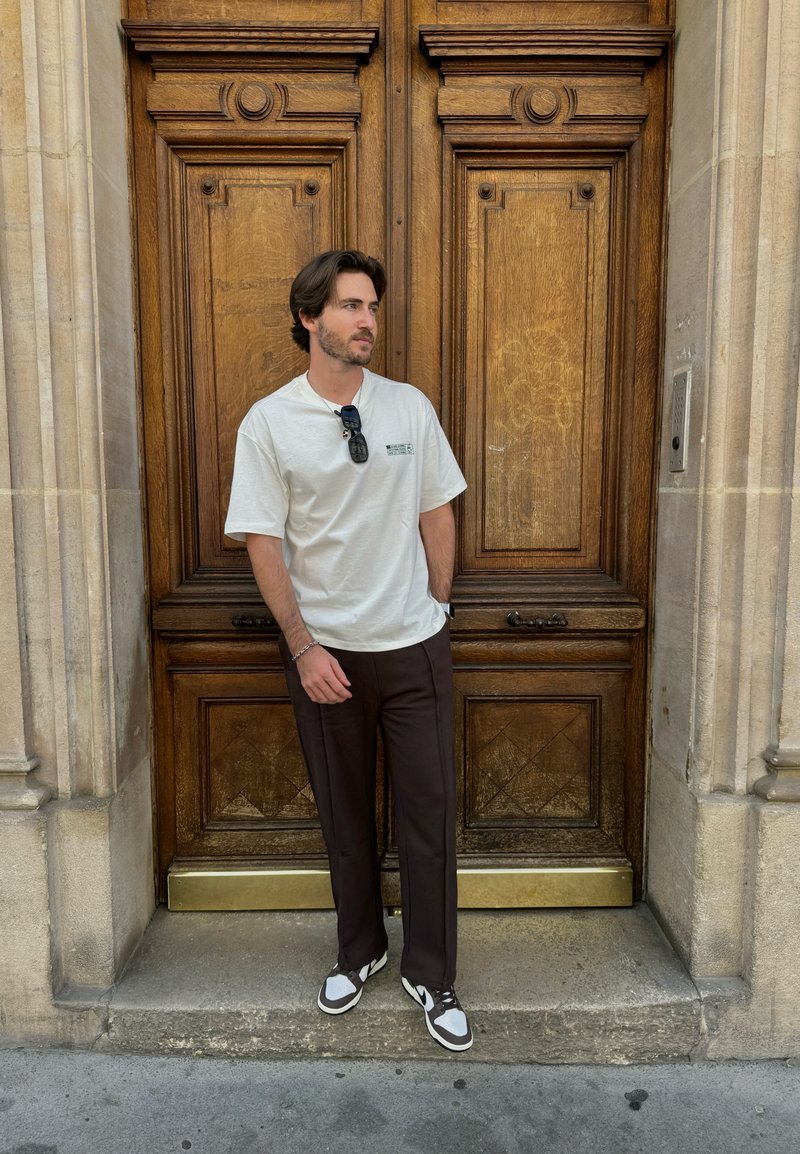 Λευκό κοντομάνικο μπλουζάκι με πράσινο λογότυπο, καφέ προσαρμοσμένα παντελόνια και μαύρα-άσπρα αθλητικά παπούτσια, ποζάροντας μπροστά από μια ξύλινη πόρτα.