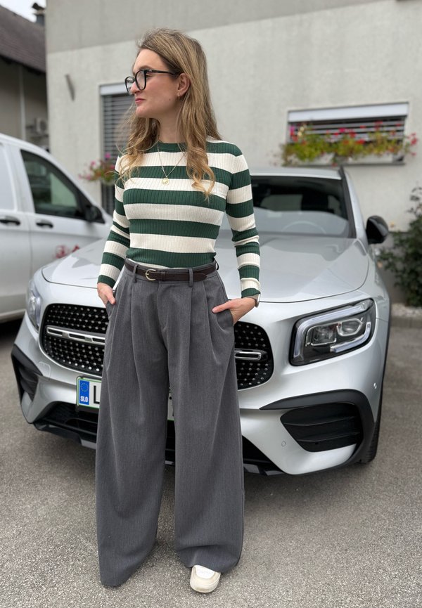 Top a righe verdi e bianche con costine, pantaloni grigi a pieghe e larghi, cintura in pelle marrone, indossato con scarpe da ginnastica bianche. Auto sullo sfondo.