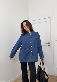 Džínová bunda v tmavě modré barvě s límečkem, dvěma předními kapsami a velkými knoflíky. Nosená přes černé kalhoty a doplněná černou taškou.