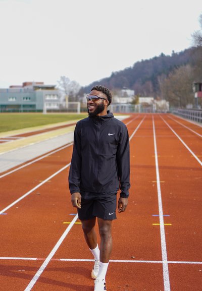 Chaqueta deportiva negra y pantalones cortos con un logo de Nike, zapatillas blancas, gafas de sol reflectantes, en una pista de atletismo roja, con un fondo difuminado.