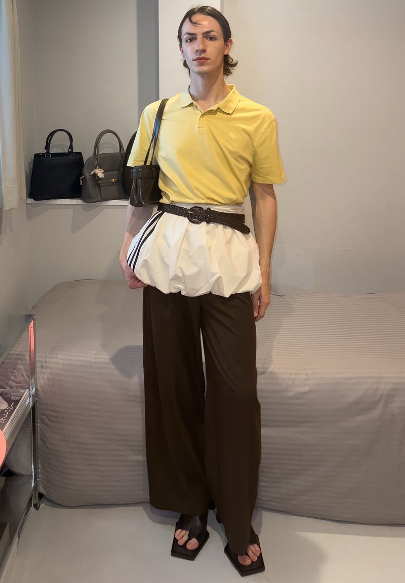 Personne portant un polo jaune, un pantalon large marron, des sandales à bout carré et un sac-ceinture blanc à rayures noires dans une pièce avec des sacs à main et un lit.