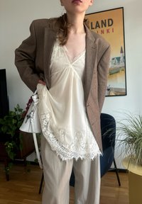 Een bruine blazer over een crèmekleurig top met kantafwerking, gecombineerd met licht gestreepte broeken. Een witte tas hangt aan de zijkant, aangevuld met kamerplanten.