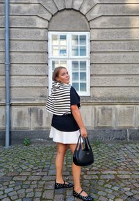 Camiseta negra, falda blanca, chal a rayas drapeado sobre los hombros. Bolsa negra con logo en forma de triángulo y flats negros con lunares. Fondo de adoquines.