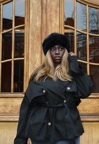 Čierny trenčkot s opaskom, veľkými gombíkmi, širokým golierom a vrstveným dizajnom; doplnený čiernou kožušinovou čiapkou. Pozadie s dreveným obkladom.