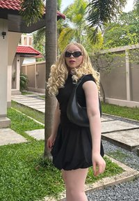 Top negru ajustat și fustă evazată, completată cu o geantă de umăr neagră. Păr lung, blond și ondulat și ochelari de soare închiși la culoare. Fundal cu iarbă verde și alee din piatră.