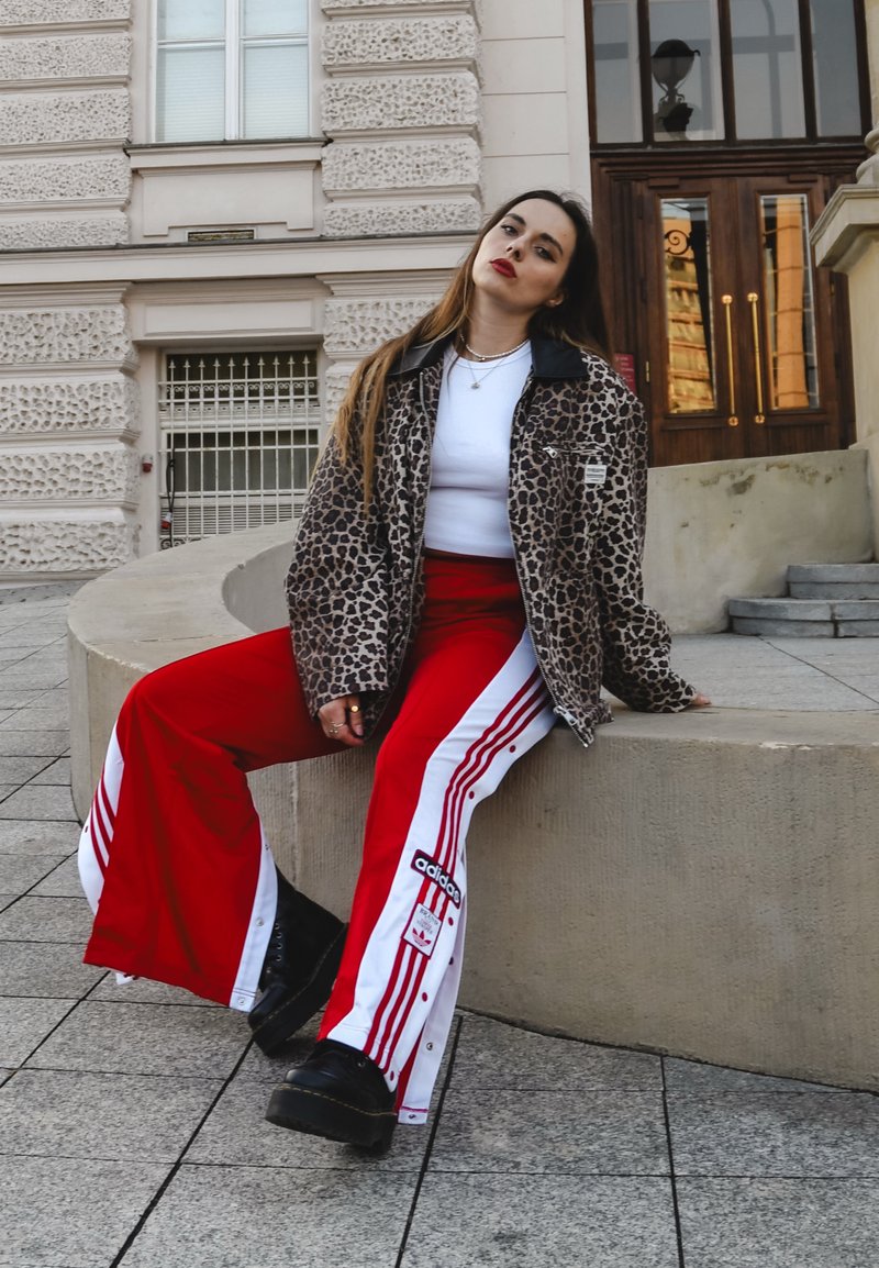 Τζάκετ με λεοπάρ σχέδιο, λευκή μπλούζα, κόκκινες φαρδύες παντελόνες με λευκές ρίγες και λογότυπο Adidas, μαύρες πλατφόρμες, καθισμένος/η σε καμπυλωτή επιφάνεια από τσιμέντο.