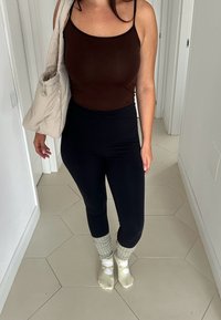 Brun ärmhållen topp, svarta högmidjade leggings, grå benvärmare över ljusa strumpor och en beige axelväska. Bakgrund med hexagonala kakelplattor.
