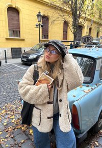 Junge Frau mit Brille und gemustertem Hut hält ein Buch, steht neben einem vintage-blauen Auto mit einem "DDR"-Schild auf einer mit Blättern bedeckten Straße.