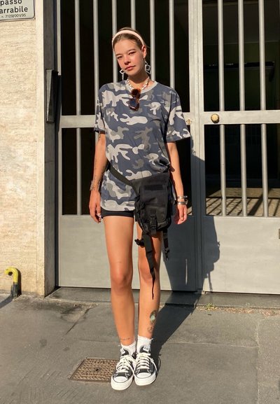 Camiseta de camuflaje en gris y azul, bolso cruzado negro, pantalones cortos negros y zapatos negros con suelas blancas. La persona está de pie frente a una puerta de metal.