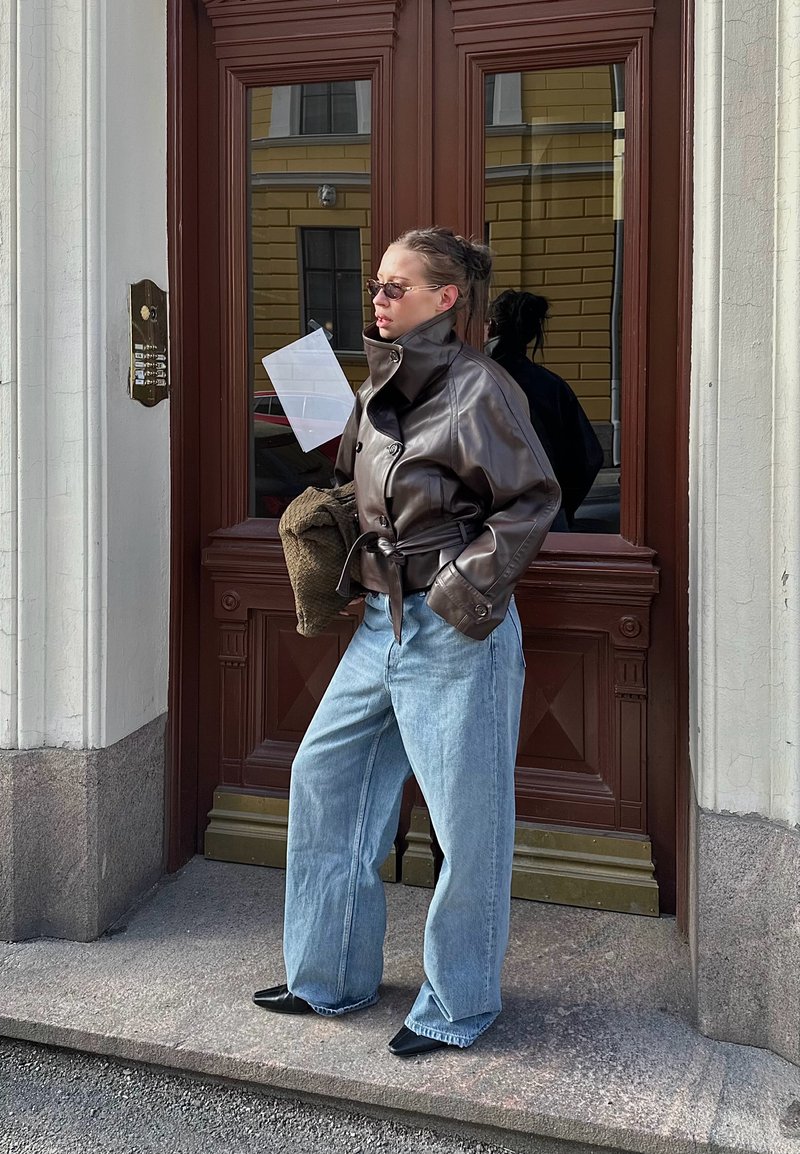 Mujer con chaqueta de cuero marrón, vaqueros de pierna ancha azul claro, zapatos negros puntiagudos y gafas de sol, parada junto a una puerta doble de madera marrón.