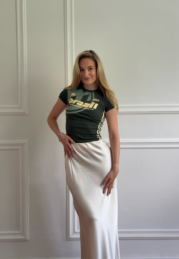 Donna con lunghi capelli biondi, indossa una t-shirt grafica verde scuro e una lunga gonna di raso bianca, in piedi contro un muro semplice con modanature decorative.
