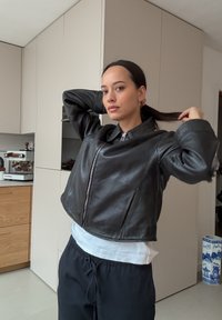 Veste courte en cuir noir avec un col montant et une fermeture éclair frontale. Portée sur une chemise blanche, associée à un pantalon décontracté. Texture lisse.