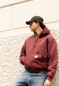 Μαρόν φούτερ με φερμουάρ, με κουκούλα που ρυθμίζεται με κορδόνι, μπροστινές τσέπες και λογότυπο. Συνδυάζεται με μπλε τζιν και μαύρο καπέλο. Υφή γκρι τοίχος στο φόντο.