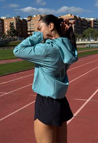 Helles türkisfarbenes, kapuzenbehaftetes Sportjackett mit lockerem Schnitt und großem schwarzem Logo auf dem Rücken, kombiniert mit schwarzen Laufshorts auf einer Laufbahn.