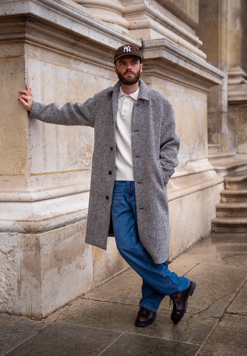 Homme portant un manteau gris à chevrons, un pull blanc, un jean bleu, des chaussures noires et une casquette marron, appuyé contre un mur en pierre près d'un escalier.