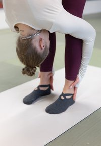 Une personne portant des chaussettes grip grises avec un design découpé, des leggings ajustés bordeaux et un haut à manches longues se penche en avant sur un tapis de yoga.