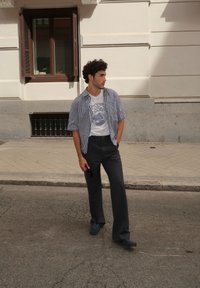 Modelka ubrana w koszulę w kratkę z krótkim rękawem na graphic t-shircie oraz szerokich, grafitowych spodniach, stoi na ulicy w pobliżu budynku.