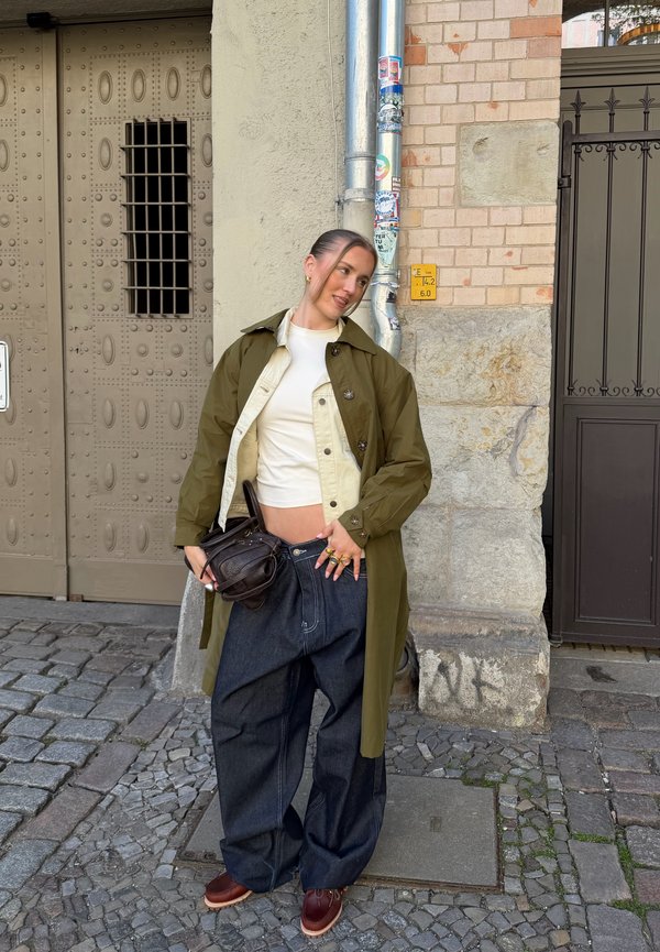 Junge Frau steht auf gepflasterter Straße, trägt grünen Trenchcoat, weißes bauchfreies Top, weite dunkle Jeans, hält eine schwarze Handtasche und lächelt.