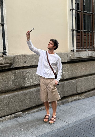 Hombre con una camisa blanca de manga larga y pantalones cortos beige sosteniendo un smartphone, vistiendo sandalias marrones, frente a una pared de piedra texturizada.