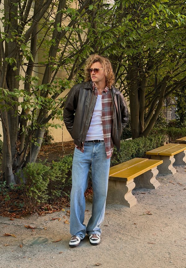 Un hombre con gafas de sol, chaqueta de cuero, camisa de cuadros, camiseta blanca y vaqueros está de pie junto a unos bancos de madera en un camino rodeado de vegetación.