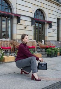Bordeaux geribbelde trui, grijze wijde broek, kastanjebruine blokhak schoenen, zwarte textuurhandtas. Buitensetting met café zitplaatsen.