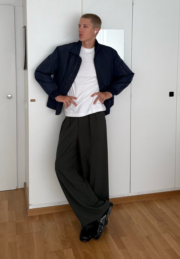 Junger Mann mit navyblauer Jacke, weissem T-Shirt, weitem schwarzen Hosen und schwarzen Schuhen, steht mit Händen in der Hüfte vor einer weissen Wand.