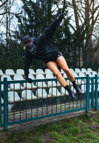Athlète portant une tenue de sport noire et un masque sautant par-dessus une clôture métallique verte dans un stade de sport extérieur vide.