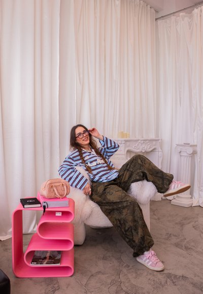 Joven mujer con camisa de rayas y pantalones de camuflaje relajándose en una silla blanca de peluche junto a una moderna mesa auxiliar rosa con libros y un bolso rosa.