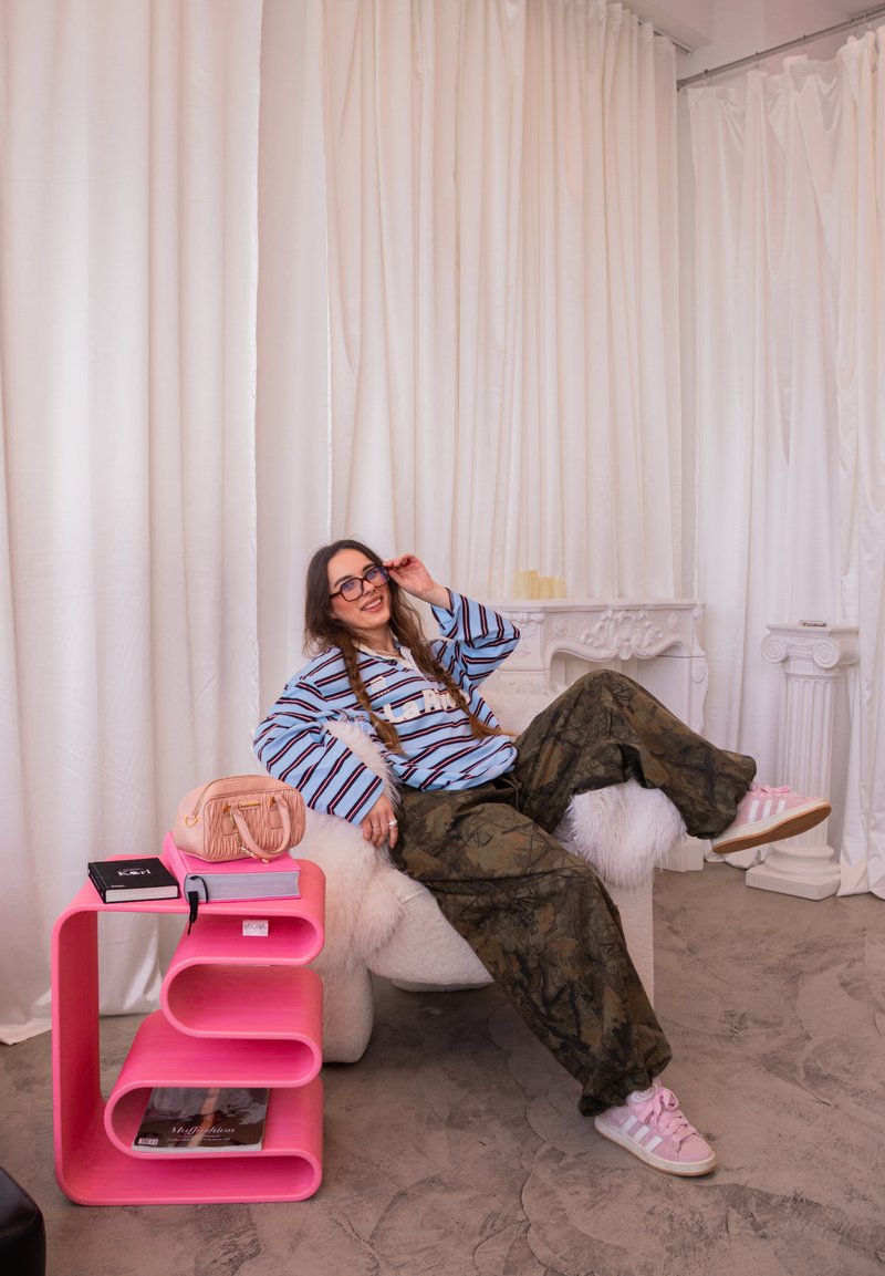Jeune femme en chemise rayée et pantalon camouflage se détendant sur un fauteuil blanc en fourrure à côté d'une table d'appoint moderne rose avec des livres et un sac à main rose.