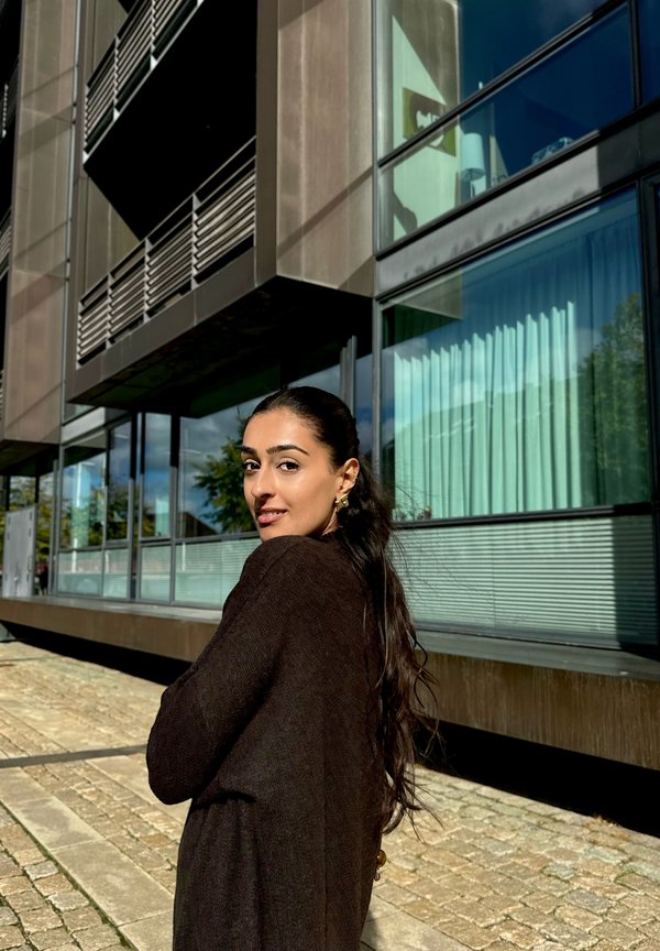 Brązowy sweter dzianinowy z długimi rękawami i luźnym krojem, stojący przed nowoczesnym budynkiem z dużymi szklanymi oknami i zielonymi firanami.