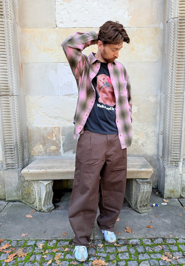 Hombre de pie al aire libre con pantalones marrones, camiseta negra con estampado gráfico y camisa rosa de cuadros, con una mano tocando la parte trasera de su cabeza.