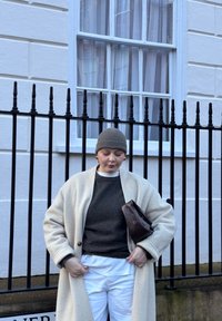Personne portant un manteau beige, un pull gris, un pantalon blanc et un bonnet gris tricoté, tenant une pochette marron, debout devant une clôture en fer noir.
