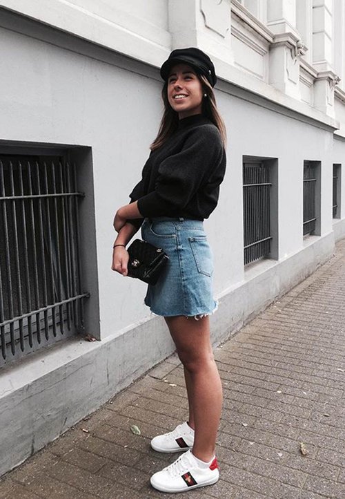 Fekete pulcsi, világoskék farmer szoknya cakkos széllel, fehér tornacipő piros részletekkel, és egy kicsi fekete kézitáska. Egy szürke falnak támaszkodva.