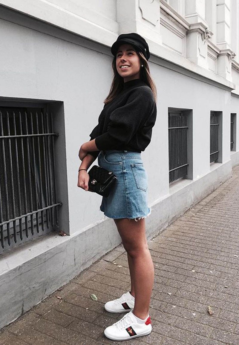 Fekete pulcsi, világoskék farmer szoknya cakkos széllel, fehér tornacipő piros részletekkel, és egy kicsi fekete kézitáska. Egy szürke falnak támaszkodva.