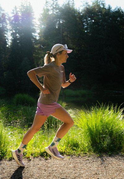 Corredor con camiseta gris y pantalones cortos morados. Gorra blanca y zapatillas con acentos rosas. Medias a rayas. Fondo de hierba verde y árboles.