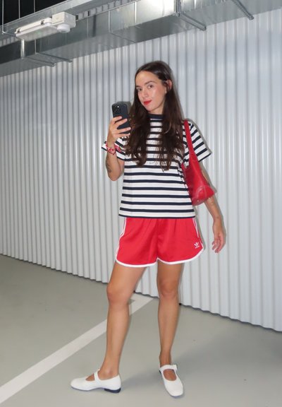 Camiseta a rayas en blanco y negro, pantalones cortos deportivos rojos con detalles en blanco, zapatos blancos, sosteniendo un teléfono, de pie en un espacio de color gris claro.