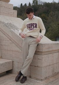 Pull beige clair avec "SUPER DRY" en bordeaux foncé, assorti à un pantalon kaki clair et des chaussures marron. L'arrière-plan présente des marches en pierre.