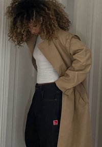 En lang, beige trenchcoat med en avslappet passform, med store knapper og sidelommer, båret over en hvit tanktopp og mørke jeans med en rød etikett.