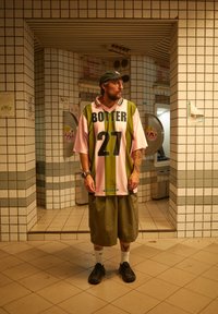 Grön och rosa oversized tröja med fet tryck av "BOLTER" och "27", matchad med olivgröna shorts och svarta skor, placerad i en kaklad korridor.
