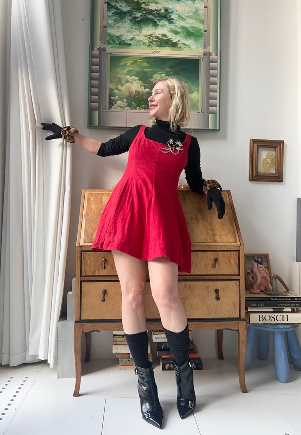 Rotes Leinenkleid mit ausgestellt Rock, schwarze Rollkragenbluse, Handschuhe mit Leopardenmuster und spitze schwarze Stiefeletten. Holztruhe und Kunstwerk im Hintergrund.