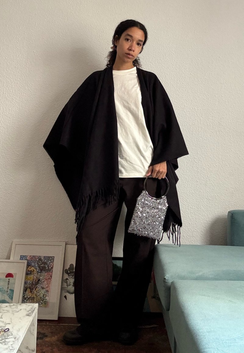 Femme debout à l'intérieur contre un mur uni portant une chemise blanche, un pantalon large noir, un châle noir, et tenant un petit sac à main argenté avec des poignées rondes.
