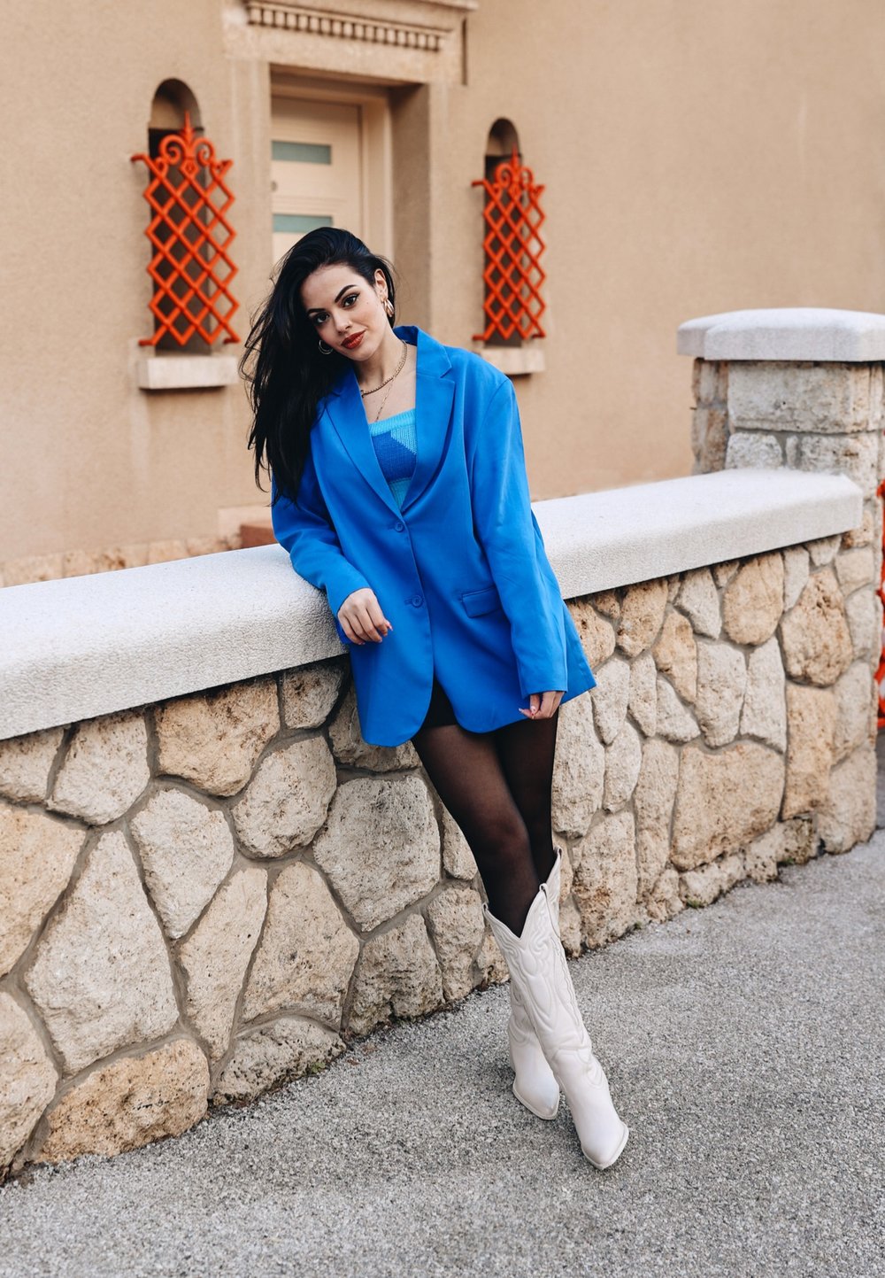 Blazer oversize bleu, superposé à un haut bleu clair. Collants noirs et bottes de cowboy blanches. Mur en pierre en arrière-plan avec des accents en treillis orange.