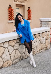 Blazer oversize bleu, superposé à un haut bleu clair. Collants noirs et bottes de cowboy blanches. Mur en pierre en arrière-plan avec des accents en treillis orange.