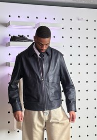 Fekete bőrdzsekit és bézs nadrágot viselő férfi egy fehér fali pegboard mellett áll, ahol cipők vannak kihelyezve a polcokon.