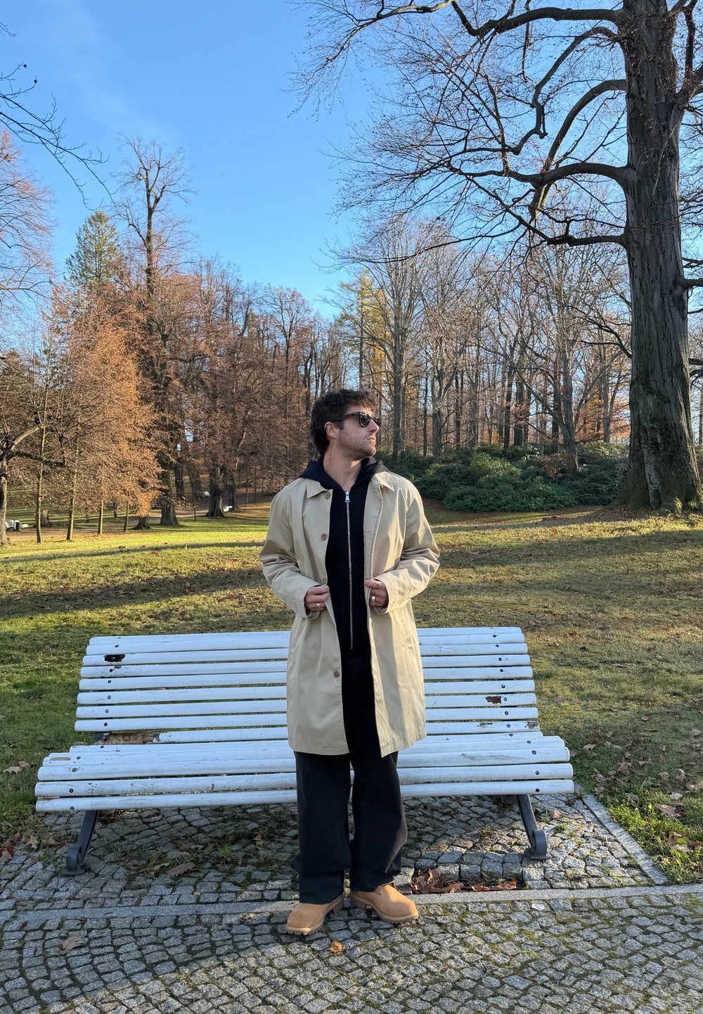 Béžový nepremokavý trenčkot so zapínaním na zips a gombíky, nosený cez čiernu mikinu s kapucňou a tmavé nohavice, doplnený o hnedé dreváky.