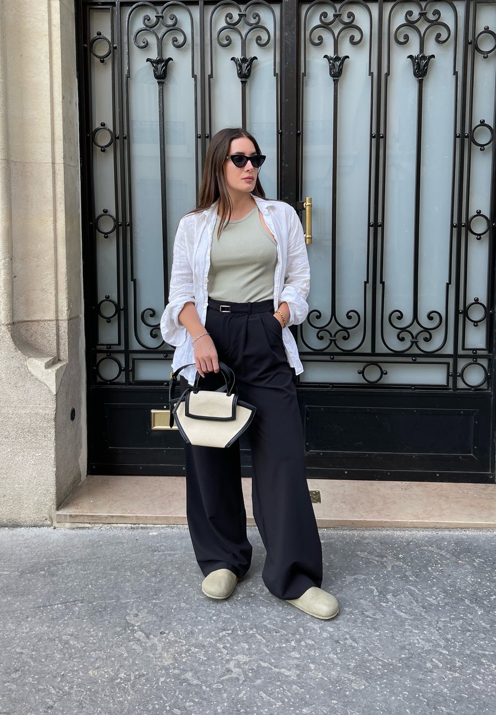 Model in een lichtgroene tanktop, een witte open shirt, zwarte wijde broek en groene instapschoenen, met een zwart en crème handtas.