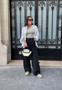 Model in een lichtgroene tanktop, een witte open shirt, zwarte wijde broek en groene instapschoenen, met een zwart en crème handtas.