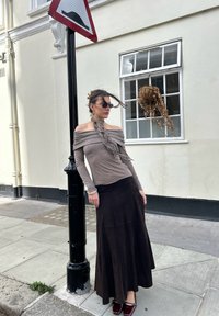 Brūns bezpiedurkņu tops ar ruffled apkakli, kombinēts ar garu, tumši brūnu zamša svārku un bordo plakanām kurpēm, fona iela.