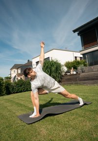 Tapete de yoga preto sobre a relva. Camisa cinza clara com padrão, calções brancos. Pessoa numa posição de lunge lateral com um braço levantado, ambiente ao ar livre.